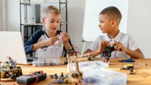Dwóch chłopców pracuje przy stole nad projektami z robotyki, używając laptopa, narzędzi i elektronicznych komponentów podczas praktycznych zajęć STEM.
